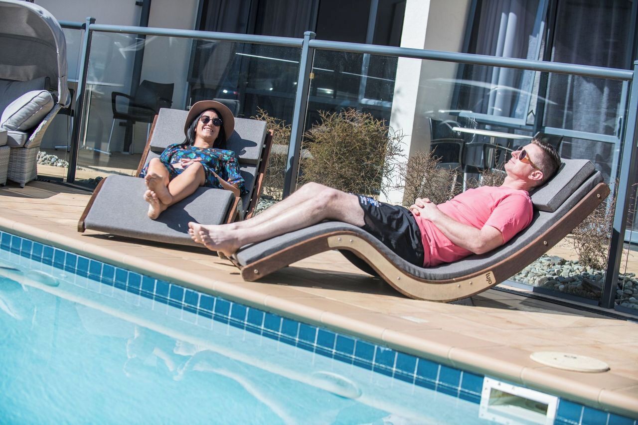 Two people relaxing on sun loungers by a poolside, with a clear view of the pool and a building in the background.