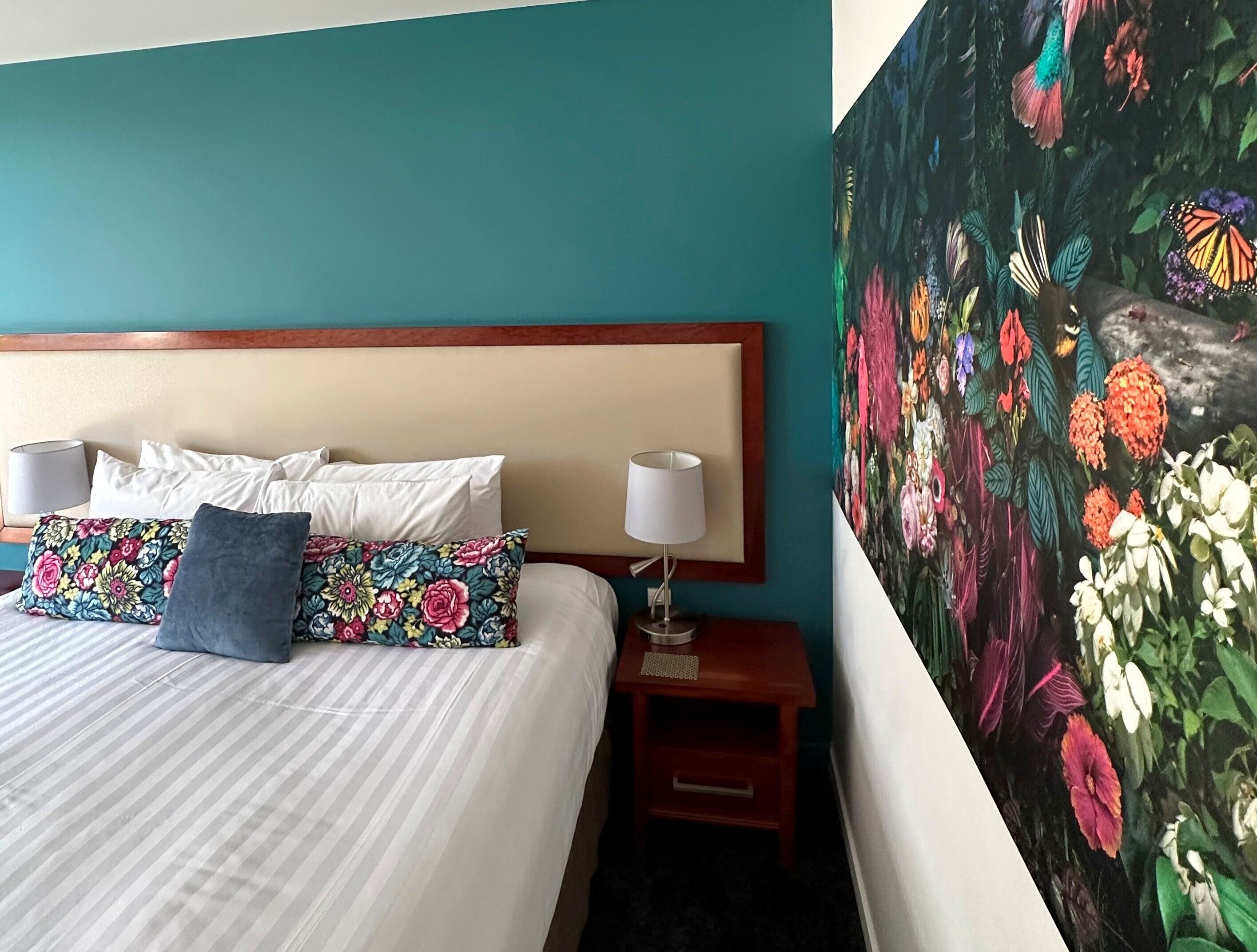 Una habitación de hotel con una cama, almohadas, una mesita de noche y una pared decorada con un mural floral.