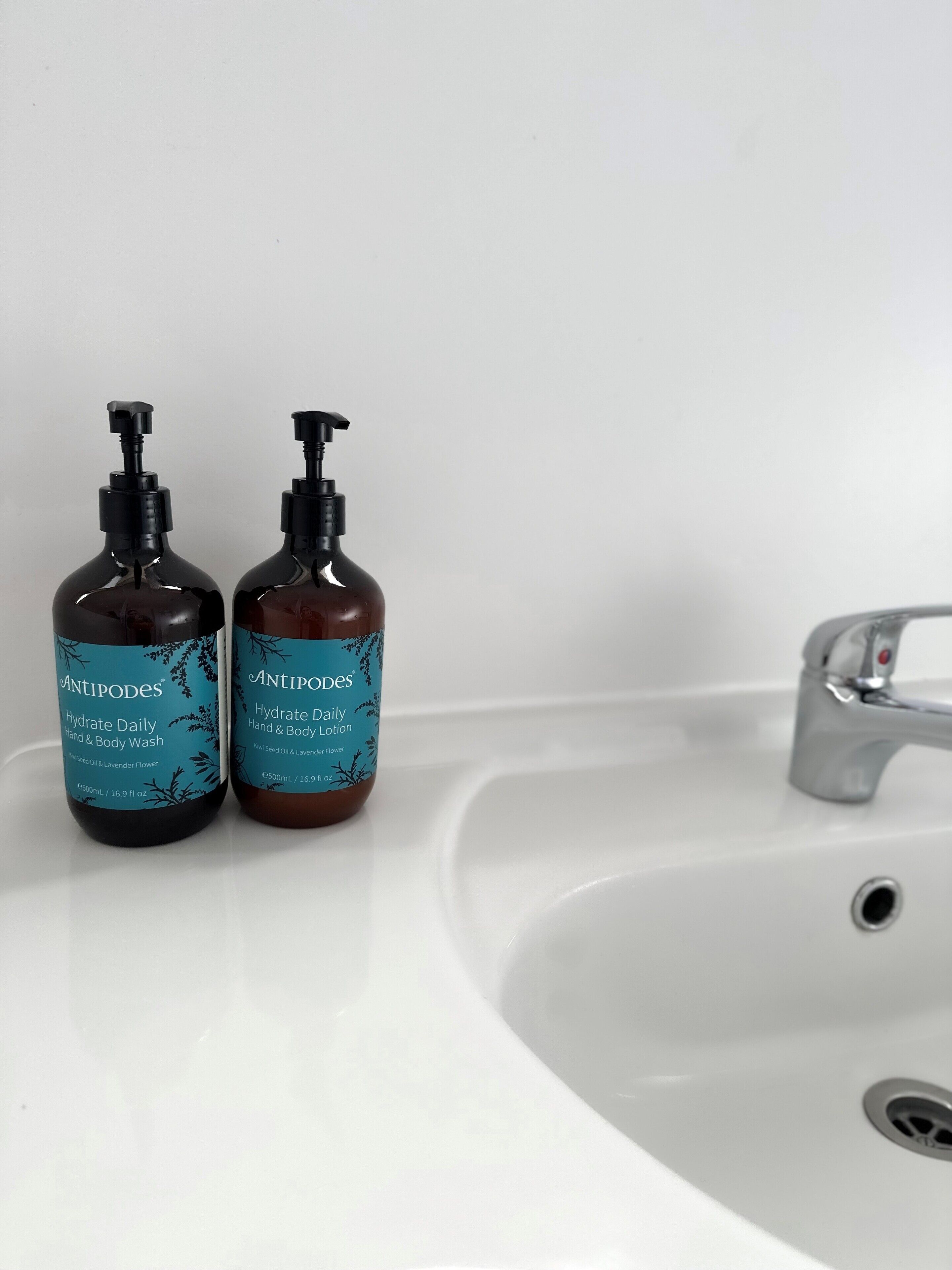 Two bottles of Antipodes Hydrate Daily Hand & Body Lotion on a white sink.
