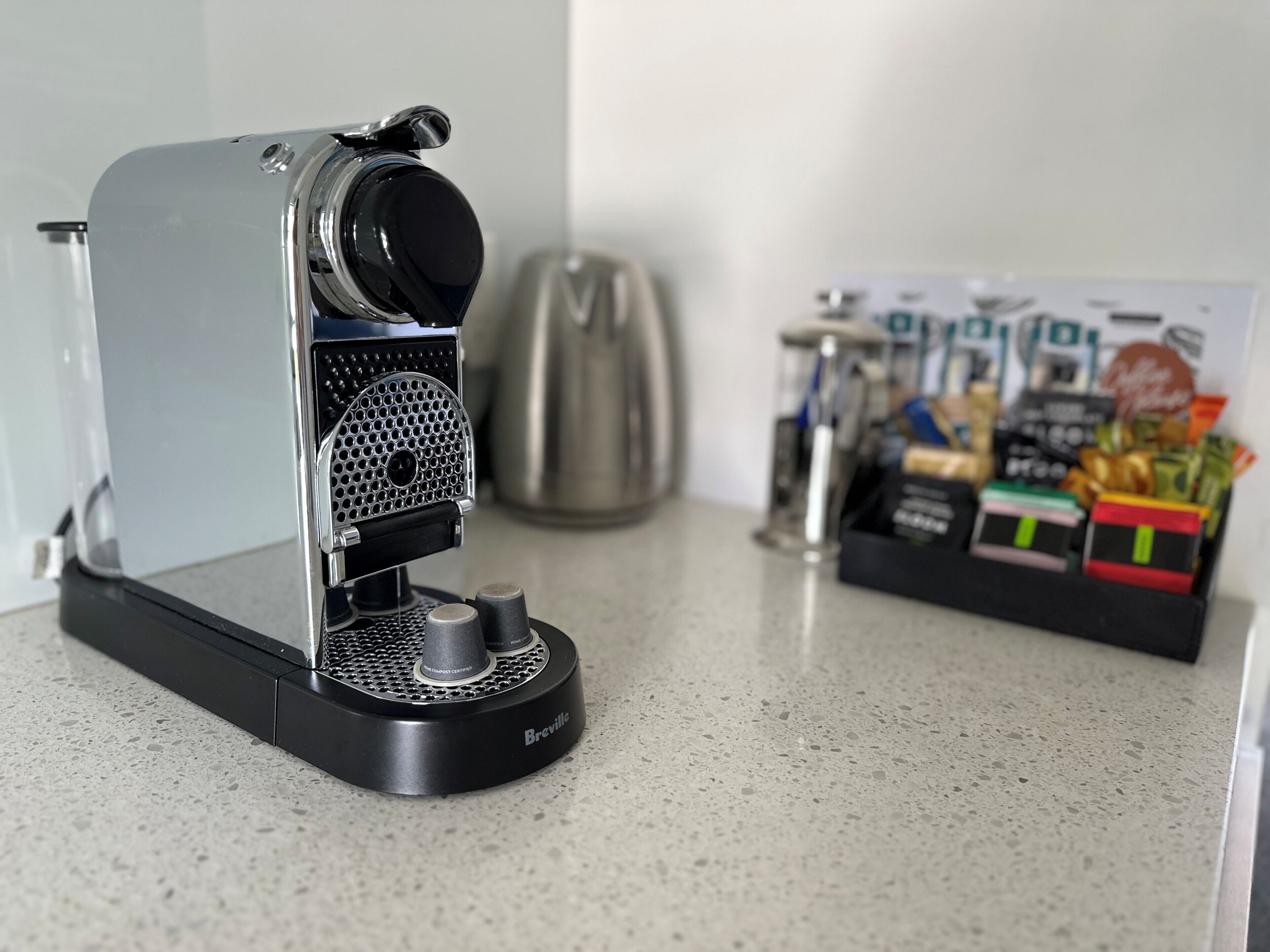 Una máquina de espresso Breville sobre una encimera de cocina, con una cafetera y una caja de cápsulas de café al fondo.