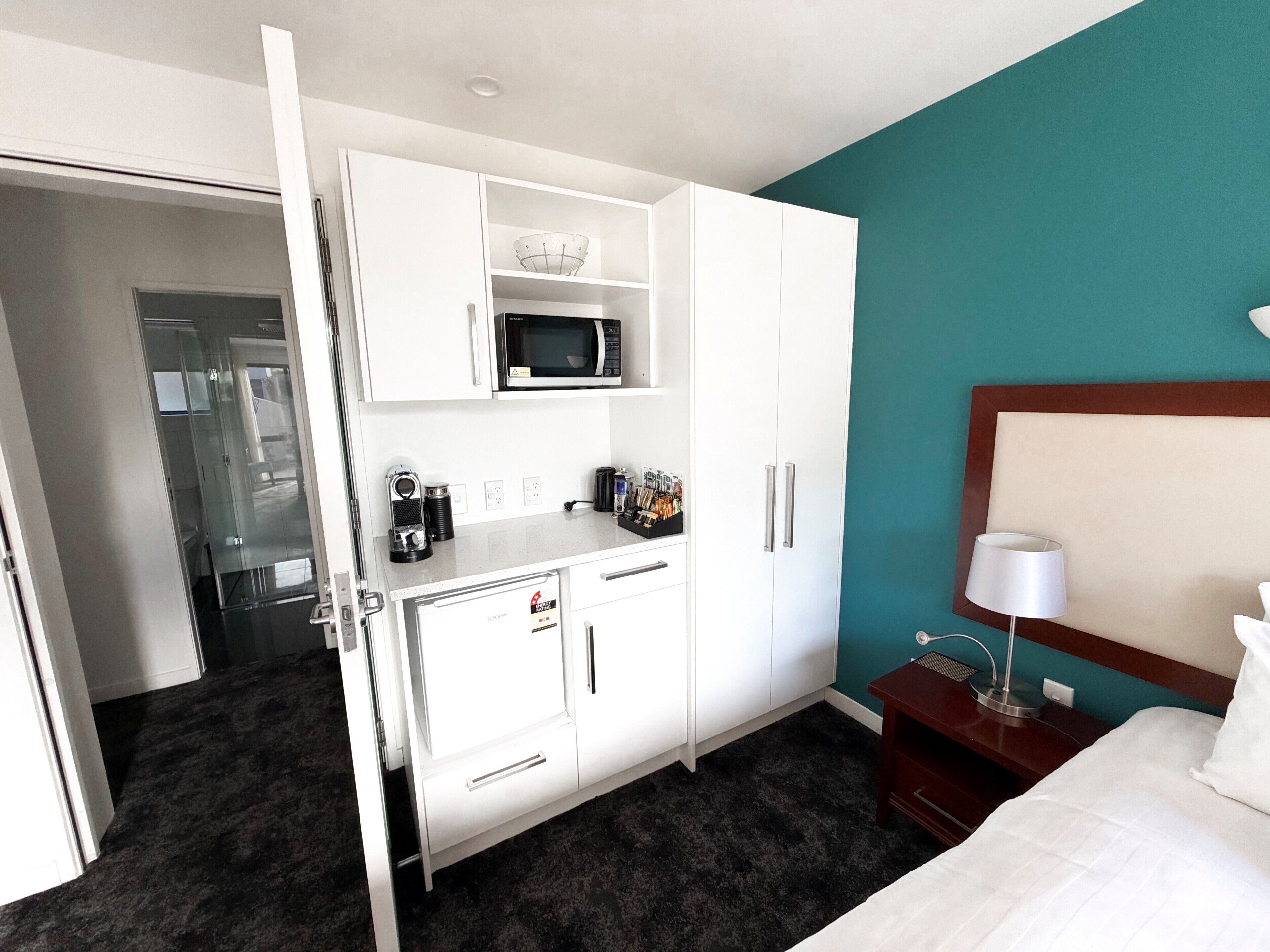 A hotel room with a kitchenette, a microwave, a coffee maker, a bedside table with a lamp, and a bed.