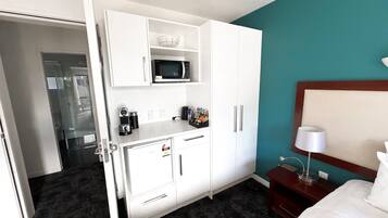A hotel room with a kitchenette, a microwave, a coffee maker, a bedside table with a lamp, and a bed.