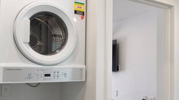 Una sala de bugaderia amb una rentadora i assecadora de càrrega frontal, una taula petita amb una cadira, i una porta que condueix a una altra habitació.