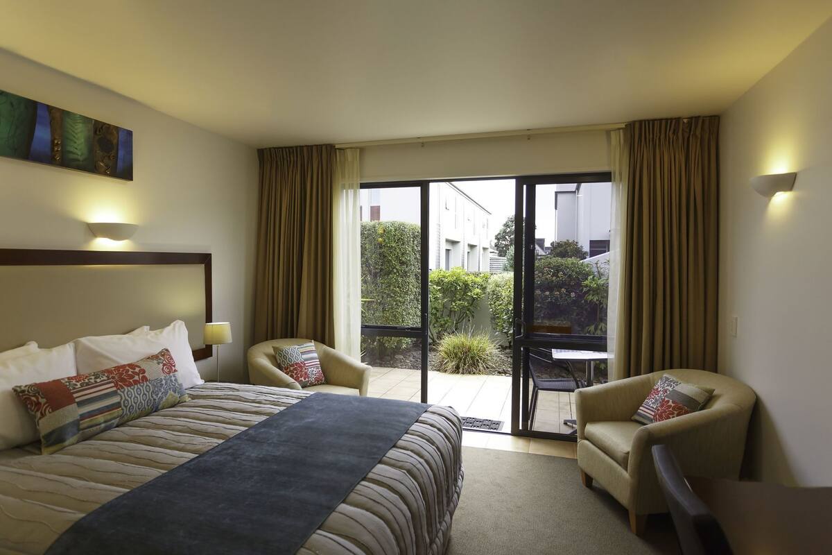 A hotel room with a bed, two armchairs, a small table, a balcony with curtains, and a painting on the wall.