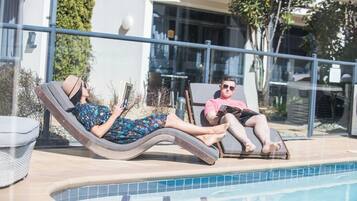 Dos personas descansando junto a una piscina, una leyendo un libro y la otra usando un teléfono inteligente.