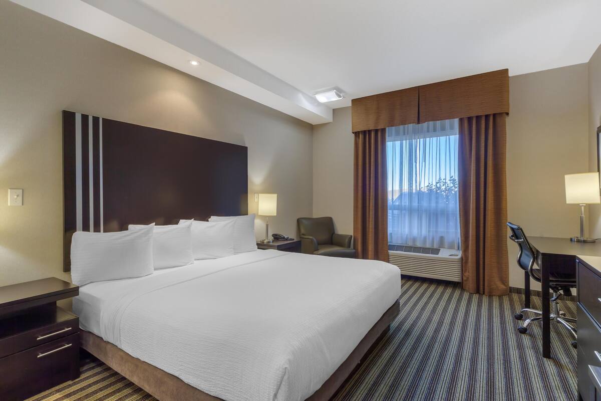A hotel room with a large bed, a desk with a chair, a window with curtains, and a wall-mounted air conditioner.