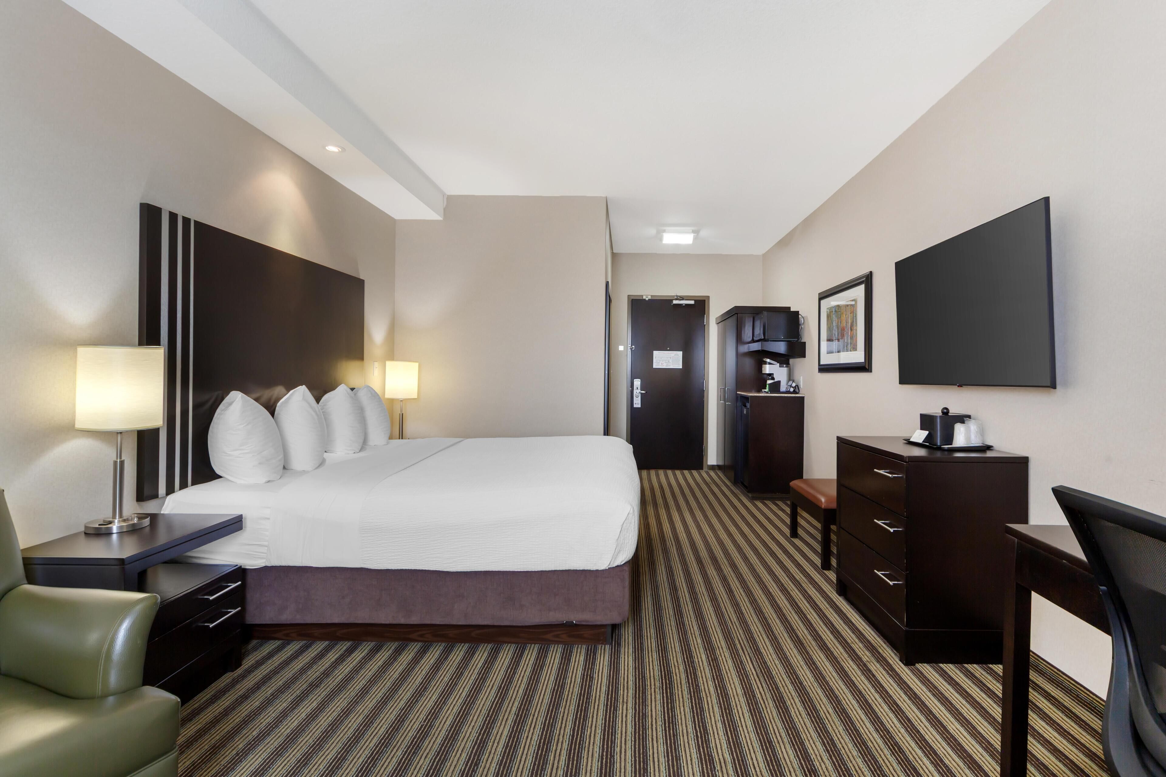 A hotel room with a large bed, bedside lamps, a desk, a chair, a dresser, a television, and a door leading to another room.