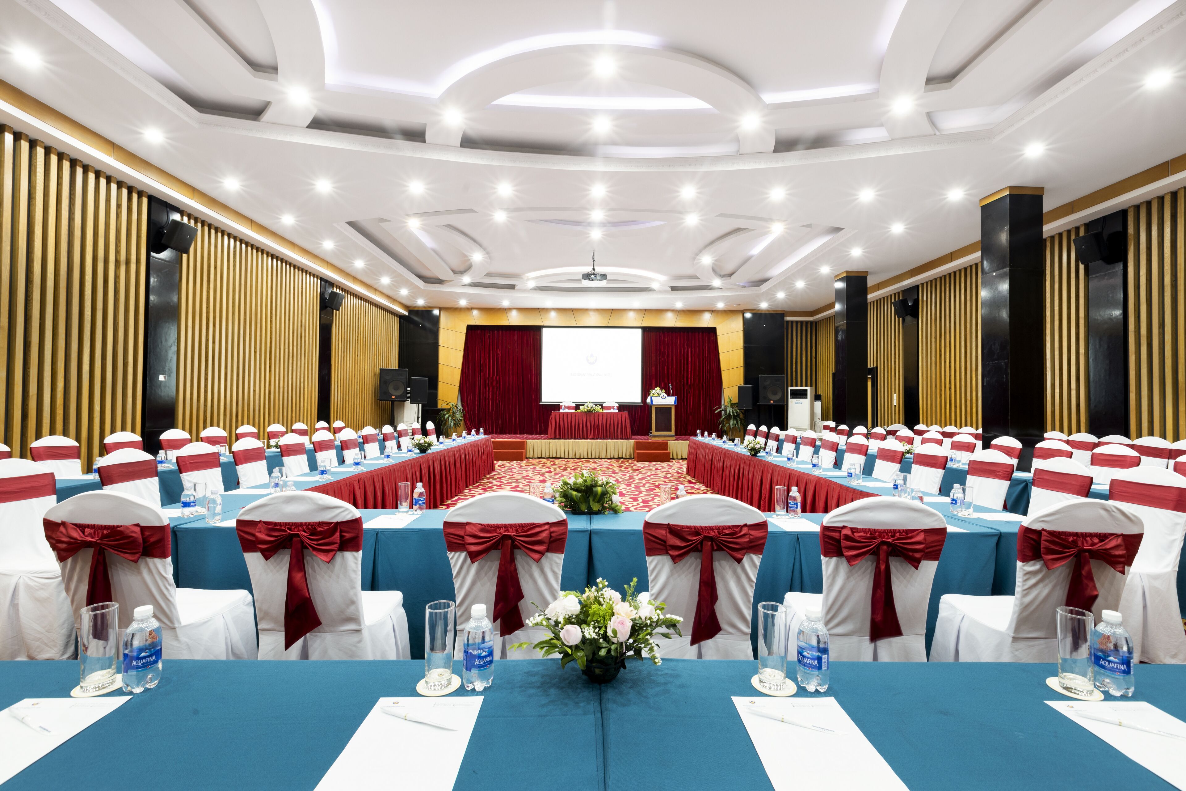 Une salle de conférence avec des rangées de tables, des chaises rouges et blanches, et une scène à l’avant.