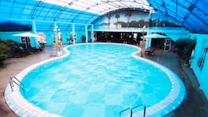 Una piscina interior con borde de azulejos azules, rodeada de sillas de descanso y sombrillas.