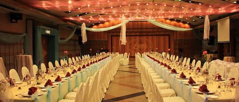 Une salle de banquet avec de longues tables dressées pour un événement formel, agrémentée de nappes et de chaises blanches, et dotée d’un plafond orné d’un éclairage décoratif.