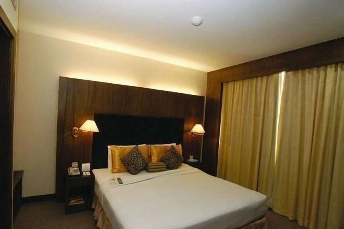 A hotel room with a large bed, two bedside lamps, a wooden headboard, and a window with curtains.