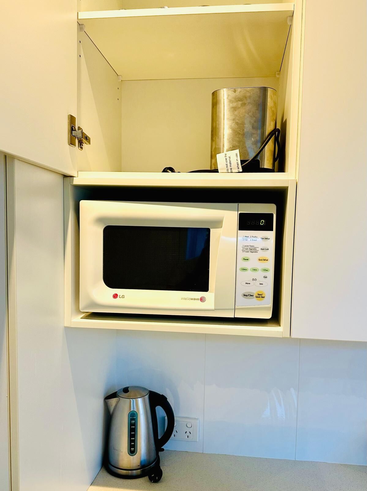 Een kleine keuken met een magnetron op een plank, een waterkoker op het aanrecht, en een roestvrijstalen bak boven de magnetron.