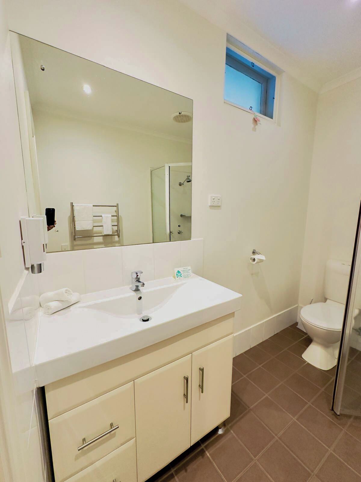 A bathroom with a white sink, mirror, and toilet. There is a towel rack and a small window above the toilet.