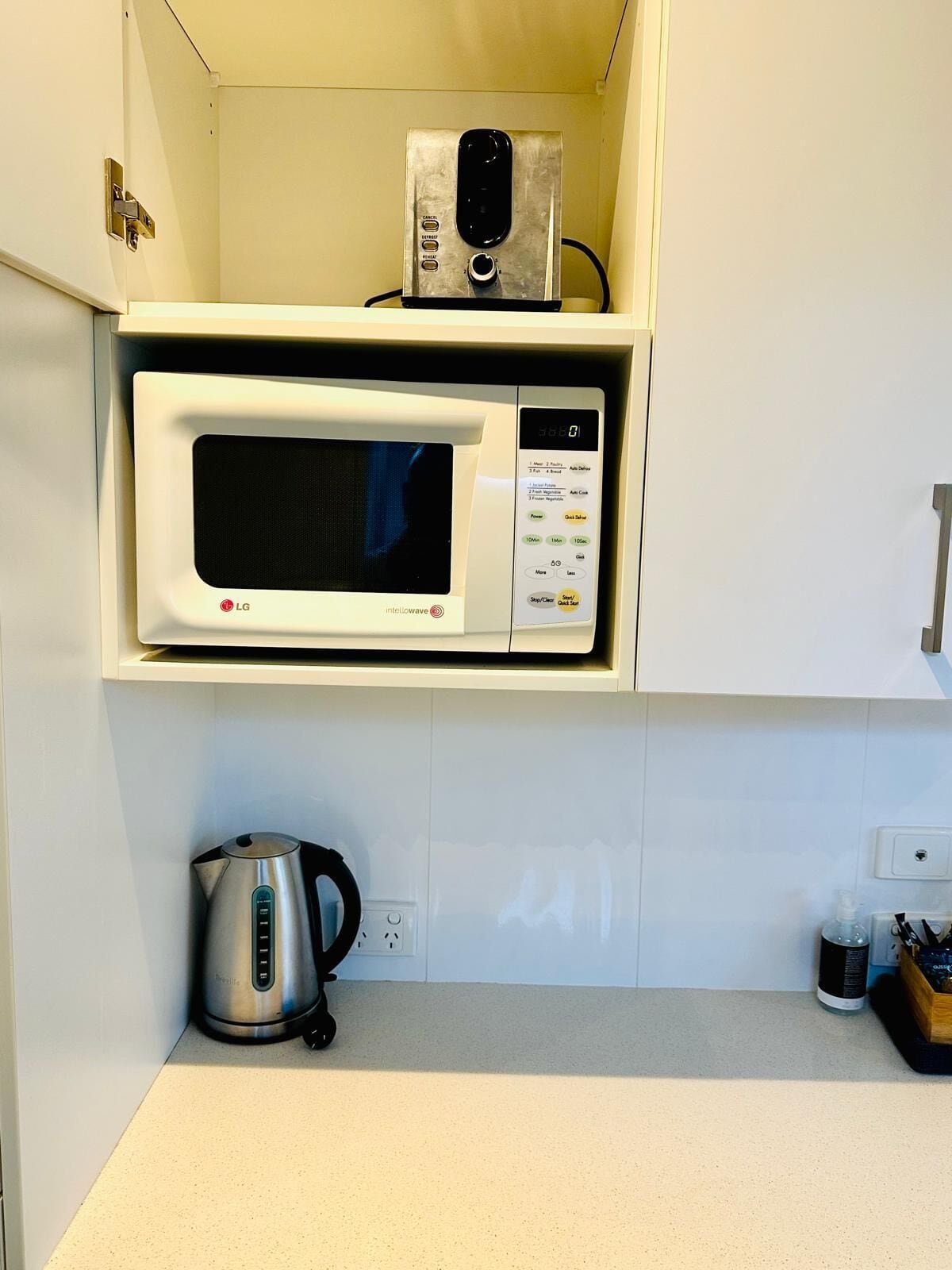 A kitchen with a microwave oven on a white shelf, a kettle on the counter, and a small cabinet with a power outlet.