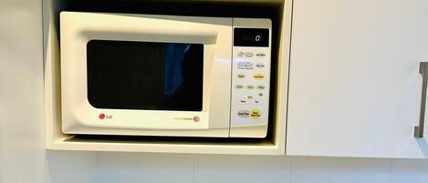 A kitchen with a microwave oven on a white shelf, a kettle on the counter, and a small cabinet with a power outlet.