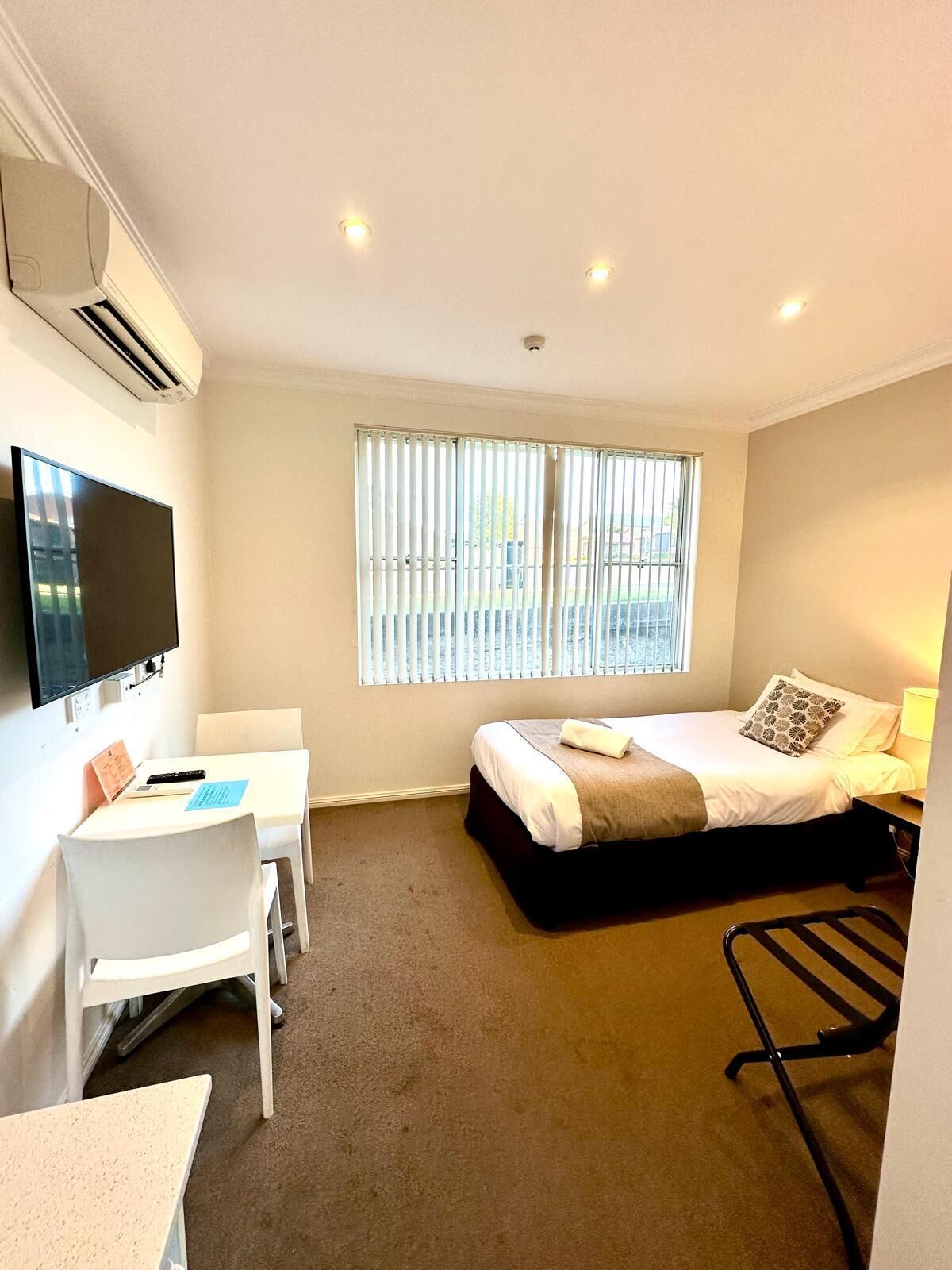A compact hotel room with a bed, a desk, a chair, a TV, and a window with blinds.
