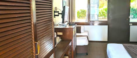 A hotel room with a bed, a desk with a chair, a television, and a window with a view of greenery outside.