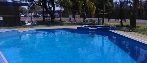 Piscina azul cristalina con una zona delimitada por una valla, rodeada de árboles y algunas sillas.
