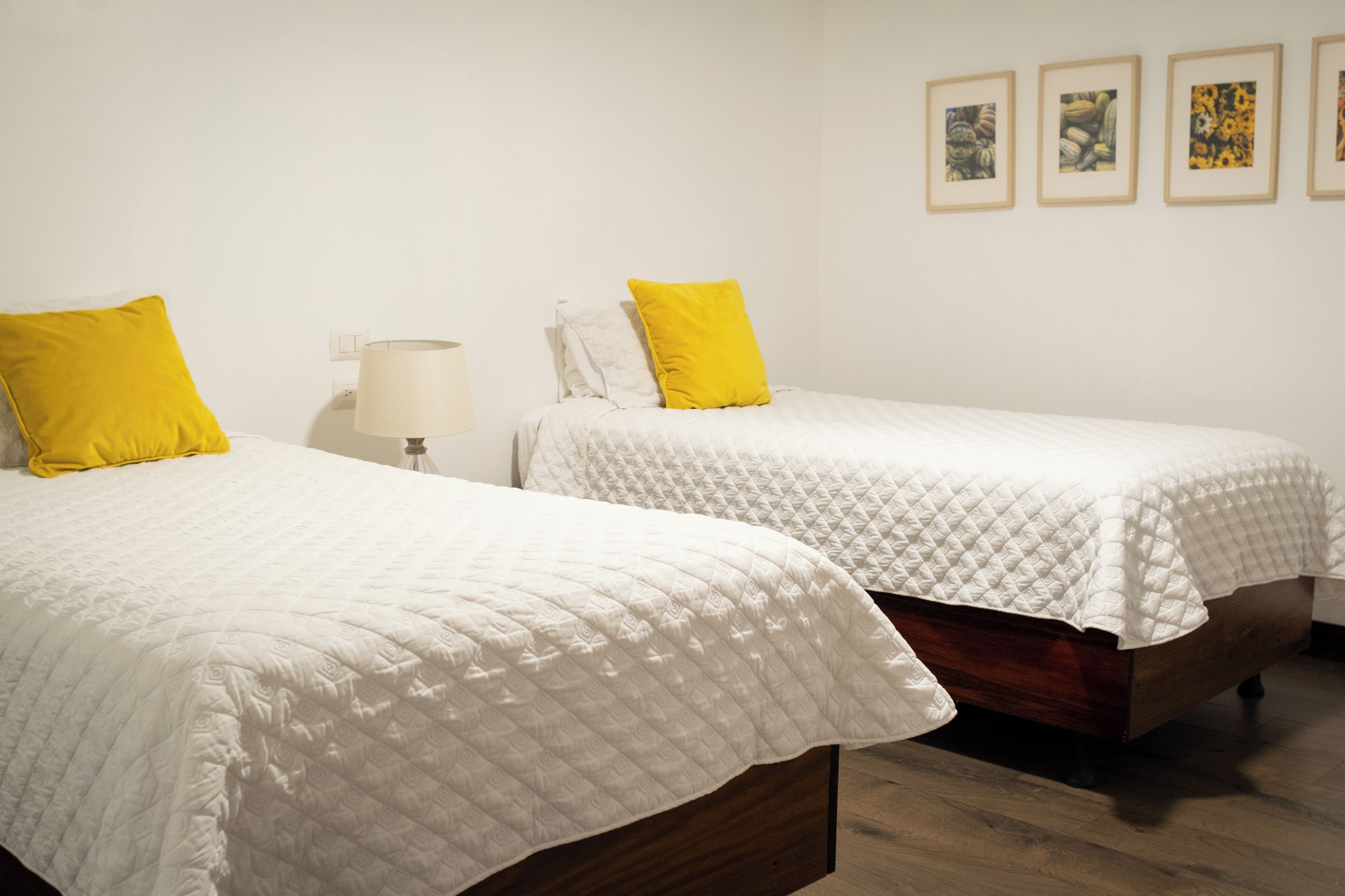Two single beds with white and yellow bedding, a bedside table with a lamp, and four framed pictures on the wall.