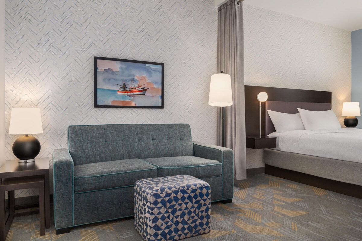 A hotel room with a grey sofa, a patterned ottoman, a bed with white bedding, and a painting of a ship on the wall.