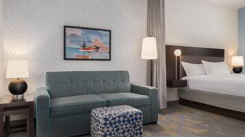 A hotel room with a grey sofa, a patterned ottoman, a bed with white bedding, and a painting of a ship on the wall.
