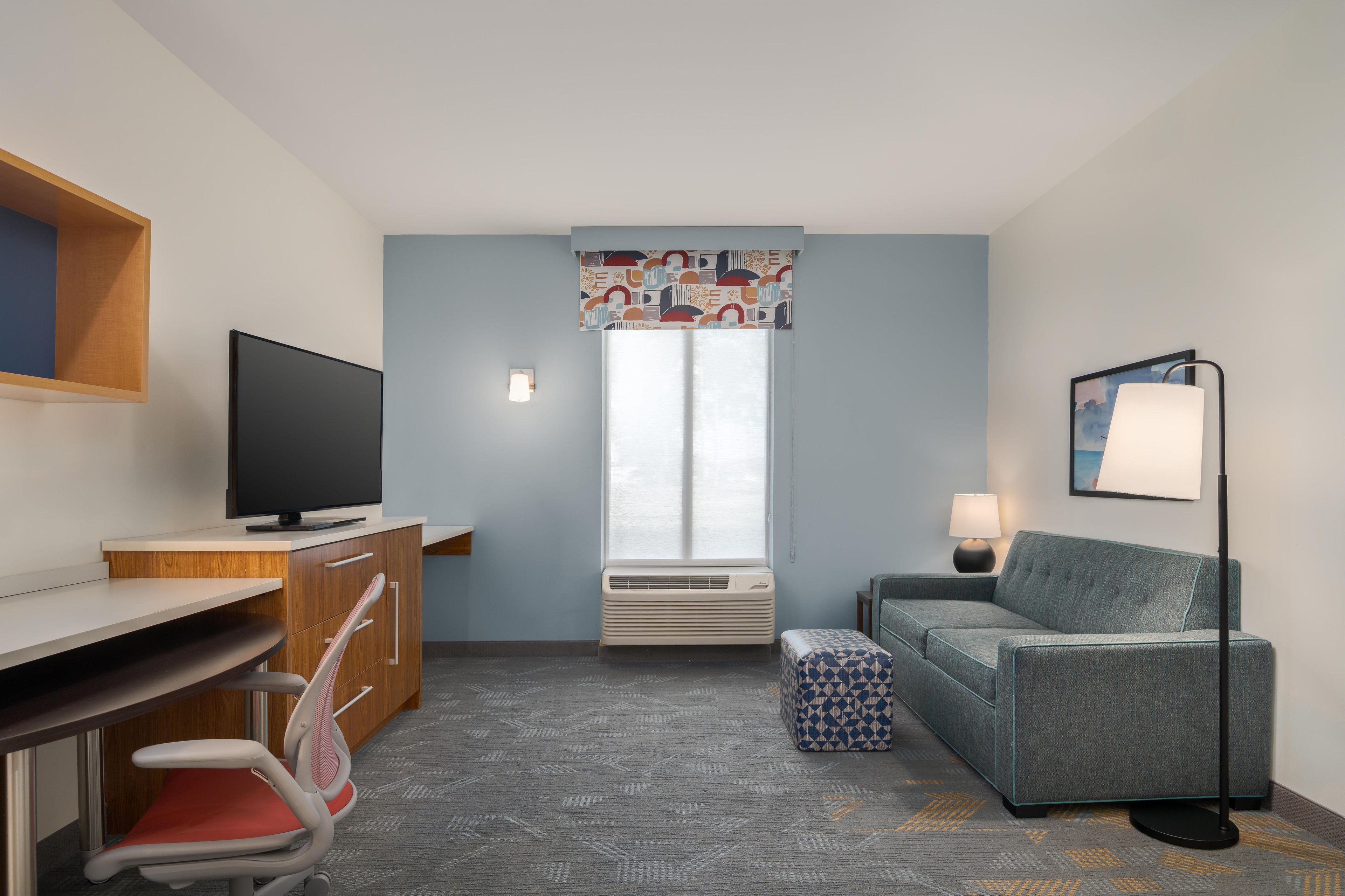 Ett hotellrum med ett skrivbord, en stol, en tv, en luftkonditioneringsanläggning, en soffa, ett soffbord, en lampa och ett fönster med mönstrade gardiner.