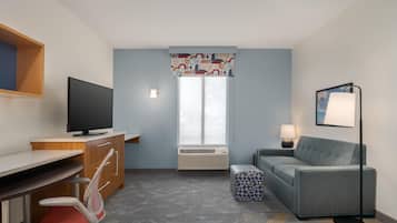 Ett hotellrum med ett skrivbord, en stol, en tv, en luftkonditioneringsanläggning, en soffa, ett soffbord, en lampa och ett fönster med mönstrade gardiner.