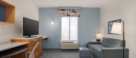 Quarto de hotel com escrivaninha, cadeira, televisão, ar condicionado, sofá, banqueta, luminária e janela com cortinas estampadas.
