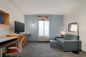 Habitación de hotel con escritorio, silla, televisor, unidad de aire acondicionado, sofá, otoman, lámpara y ventana con cortinas estampadas.