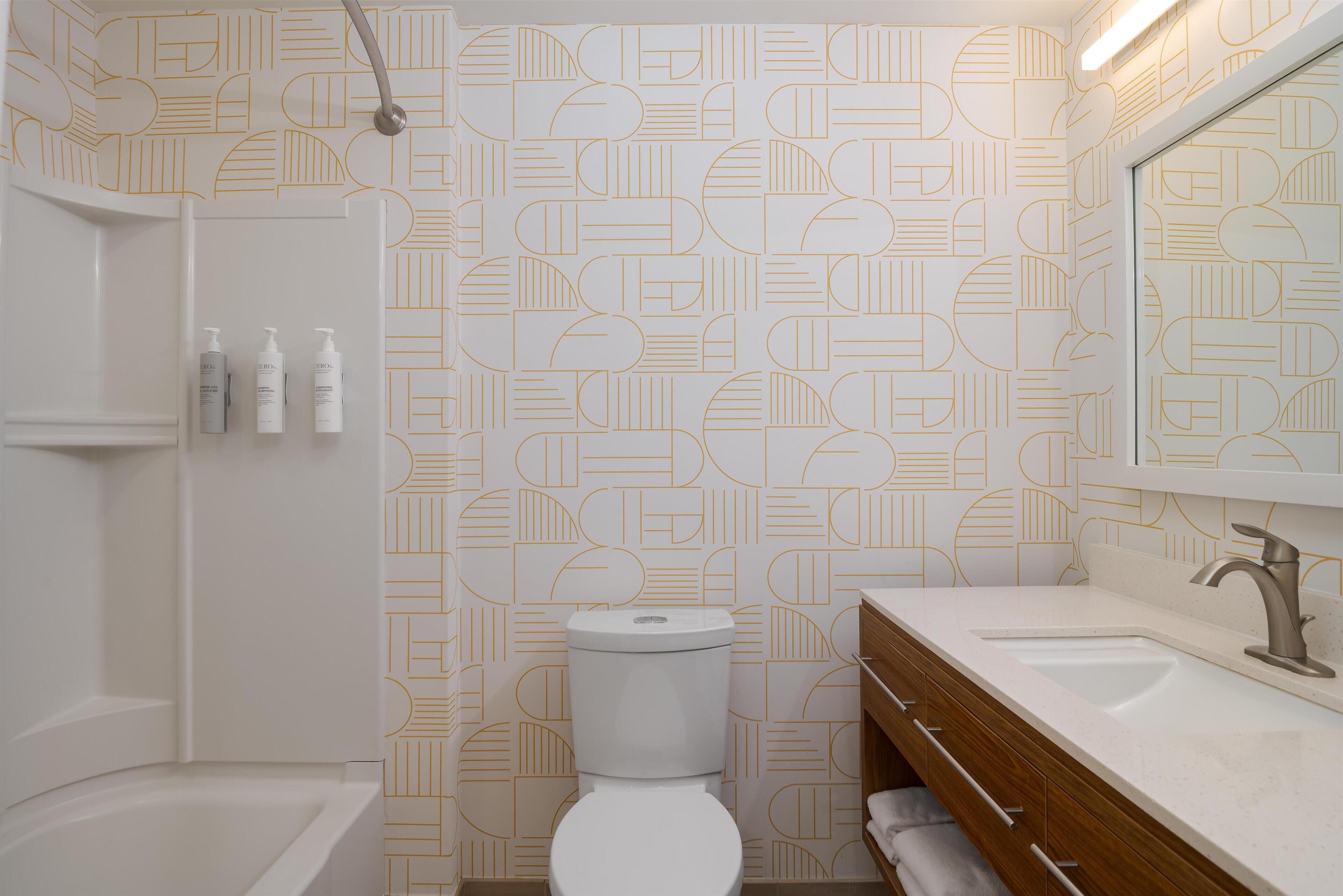 Un baño moderno con un muro de patrón geométrico, un inodoro blanco, un tocador de madera con lavamanos y una zona de ducha.