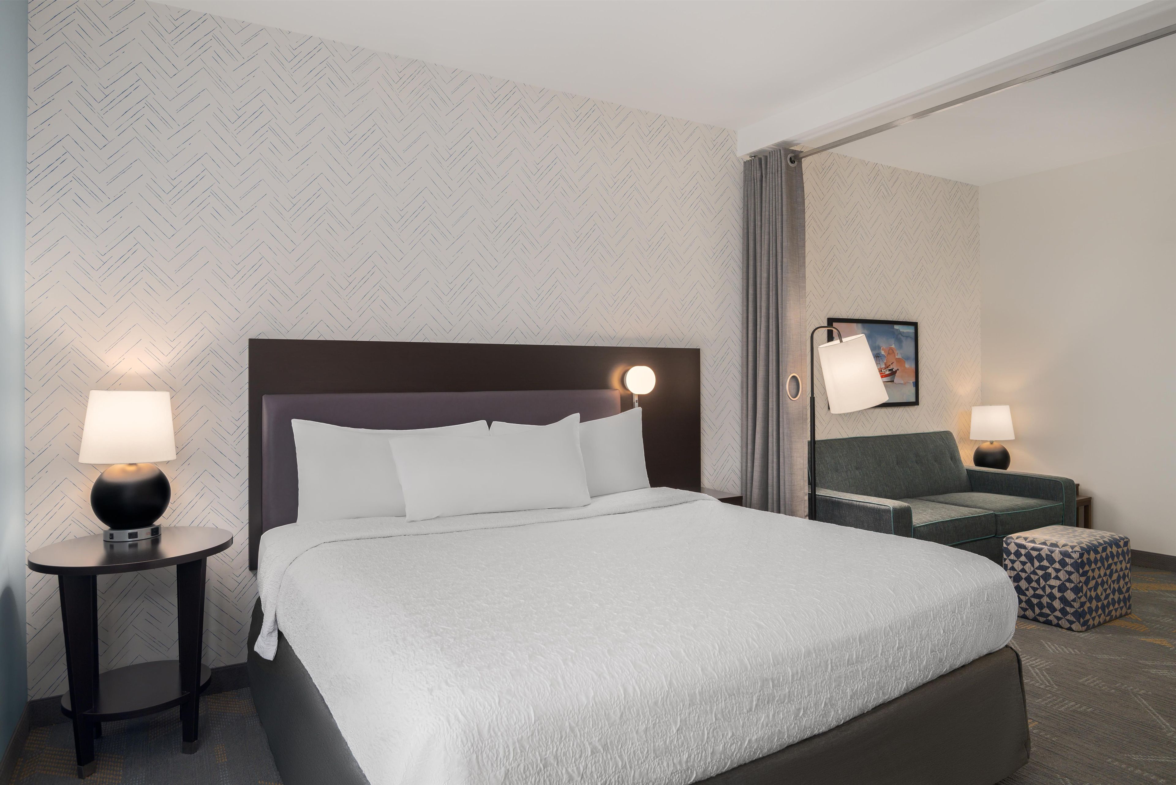 A hotel room with a large bed, two bedside tables with lamps, a sofa, and a patterned wall.