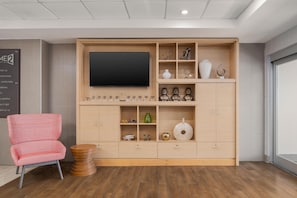 A modern living room with a large flat-screen TV, wooden shelving with decorative items, a red patterned chair, and a round wooden coffee table. - Home2 Suites by Hilton Biloxi North/D'Iberville (D'Iberville)