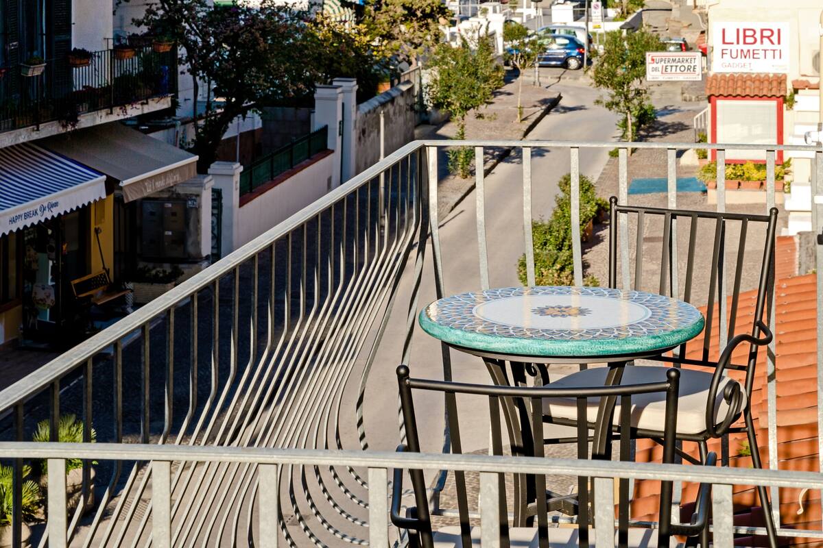 A balcony with a table and chairs overlooking a coastal street with buildings and a sailboat in the distance.