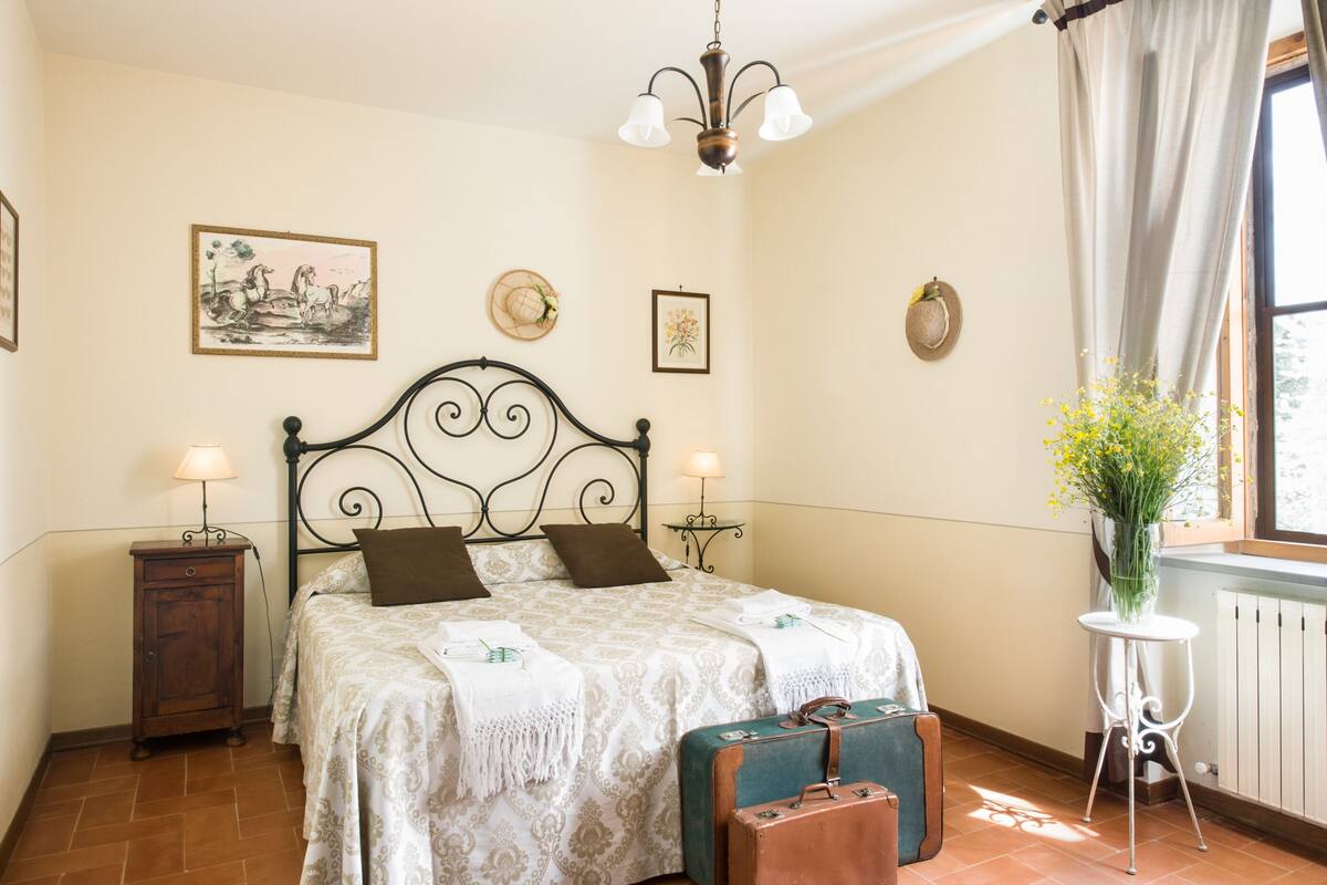 A neatly arranged bedroom with a bed, bedside tables, a suitcase, and a window with curtains.