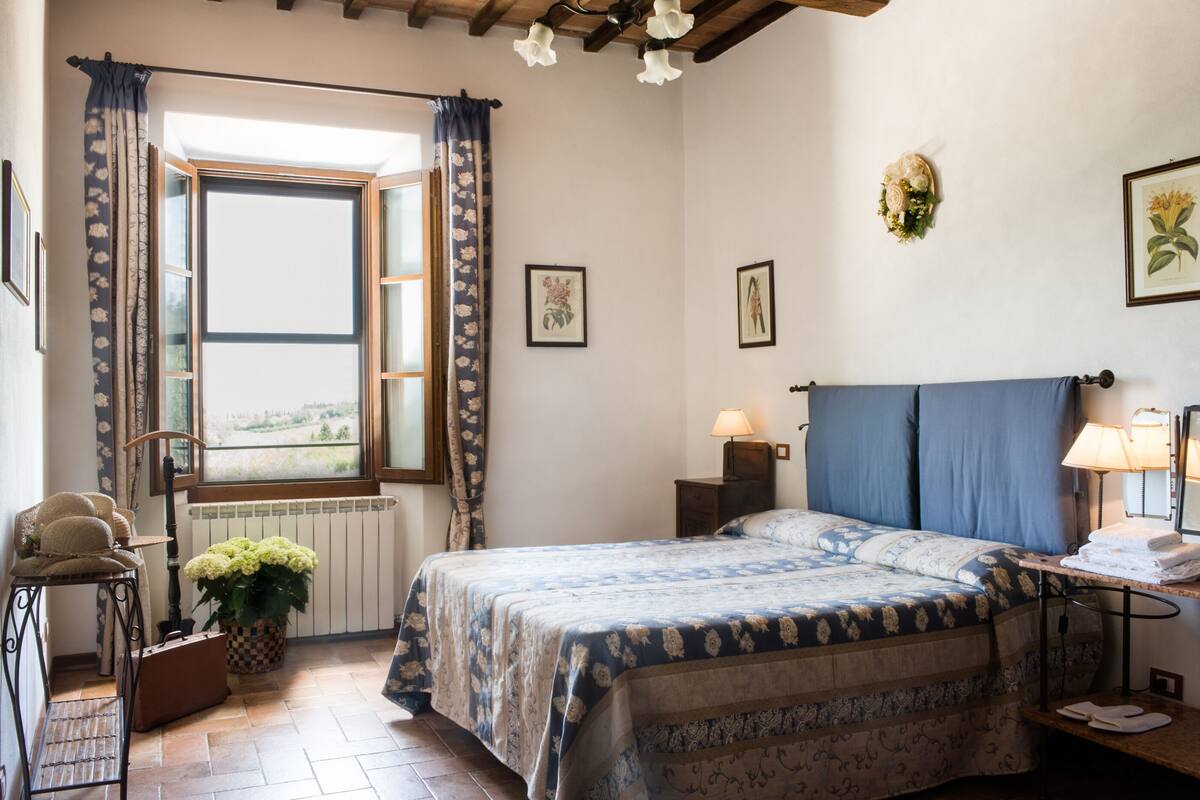 A bedroom with a bed, a window with curtains, a radiator, a bedside table with a lamp, and a wall with framed pictures.