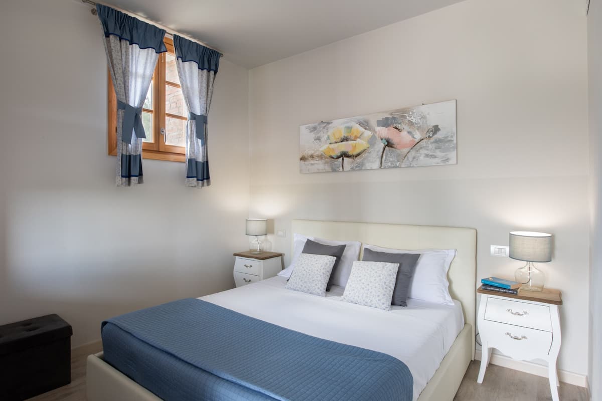 A neatly made bed with a blue bedspread, two bedside tables with lamps, a window with curtains, and a painting on the wall.