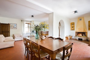 A dining area with a wooden table, chairs, and a vase of flowers. There is a fireplace and a sofa in the background. - I Pianelli (Murlo)