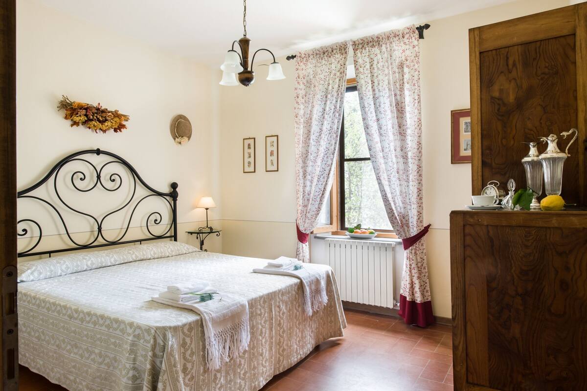 A neatly made bed with a patterned bedspread, a bedside lamp, and a window with floral curtains.