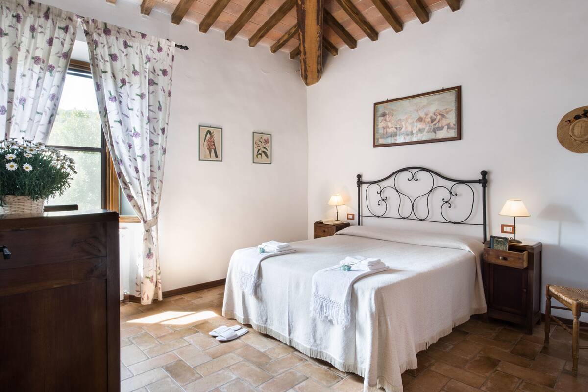 A bedroom with a bed, two bedside tables with lamps, a wooden headboard, a window with curtains, a wooden dresser, and a wall-mounted picture.