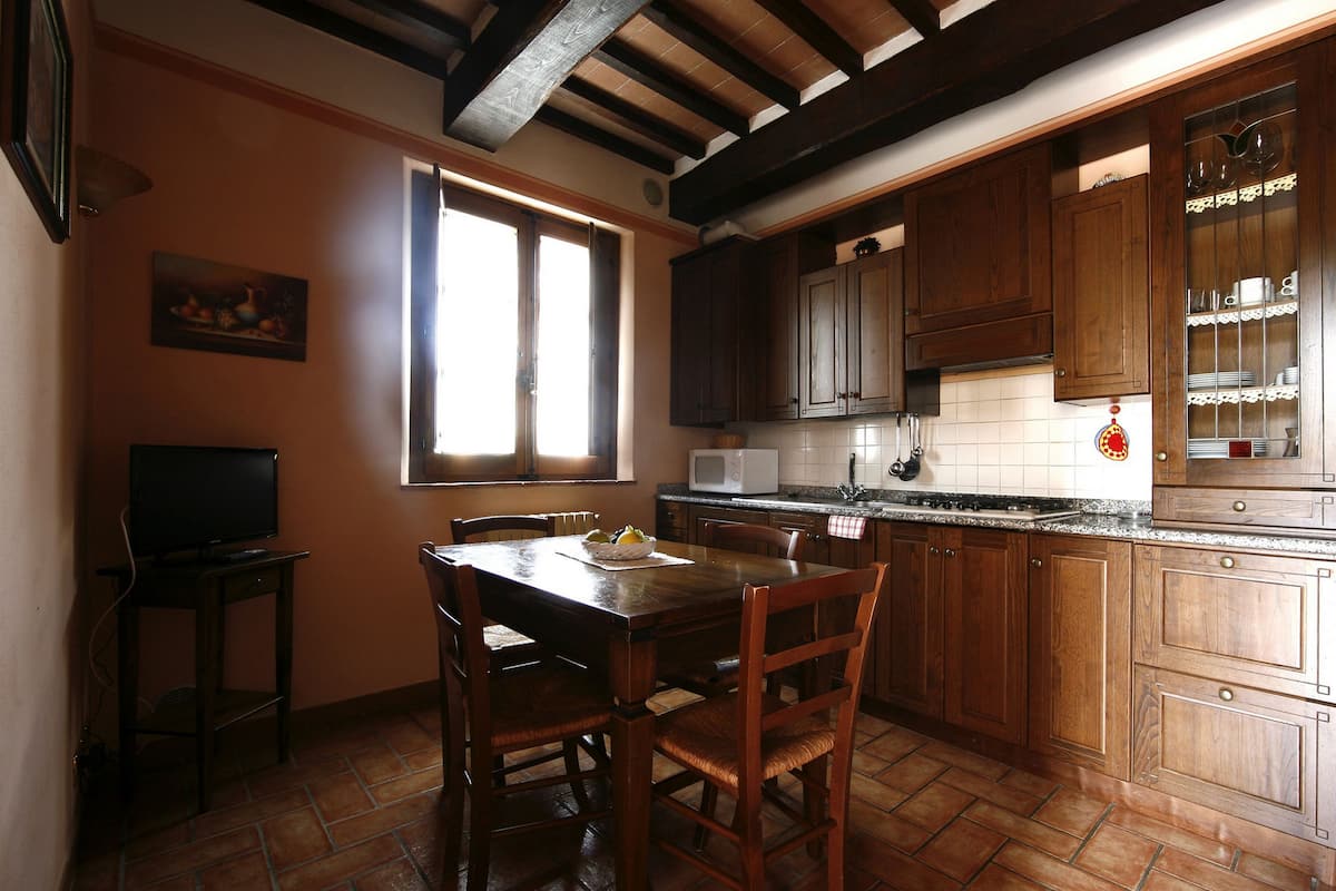 Une cuisine avec des meubles de rangement en bois, une table à manger, des chaises, un four à micro-ondes et une fenêtre avec des stores.