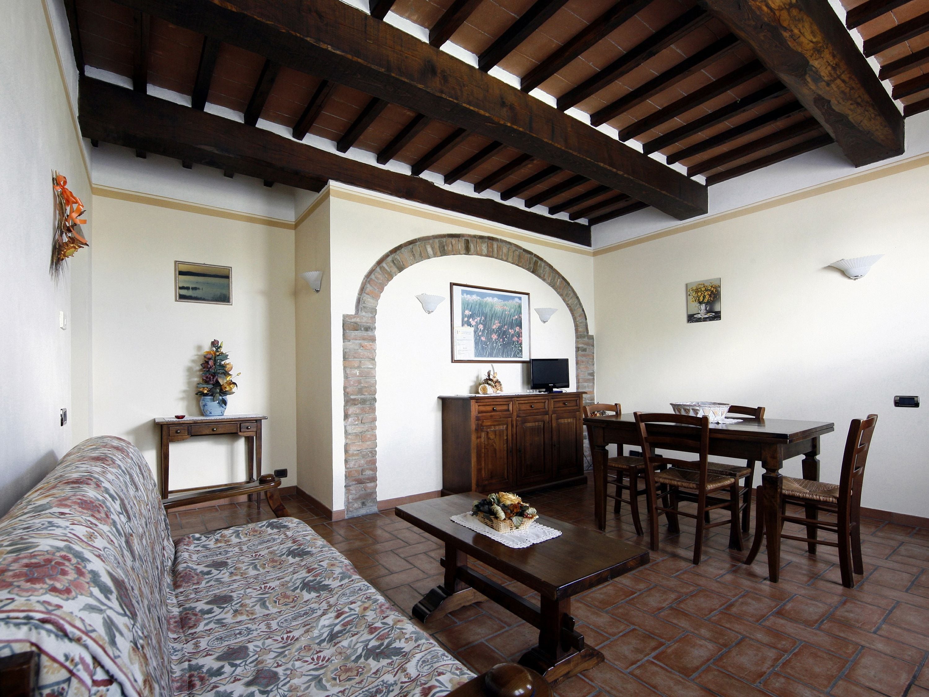 A living room with a sofa, a coffee table, a dining table with chairs, a fireplace, and a kitchen area with a brick archway.