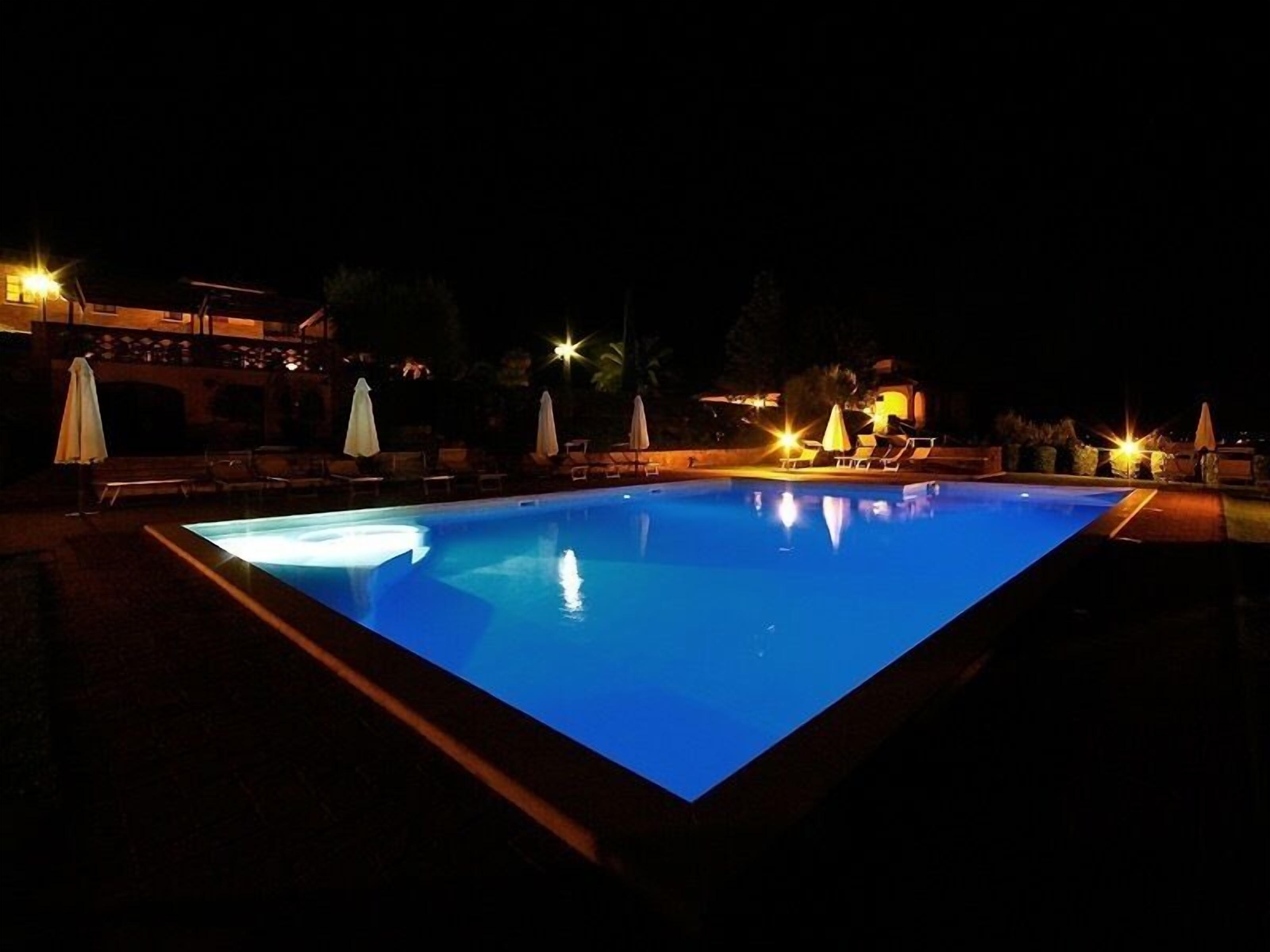 Una piscina iluminada de noche, con sillones de descanso y sombrillas alrededor.