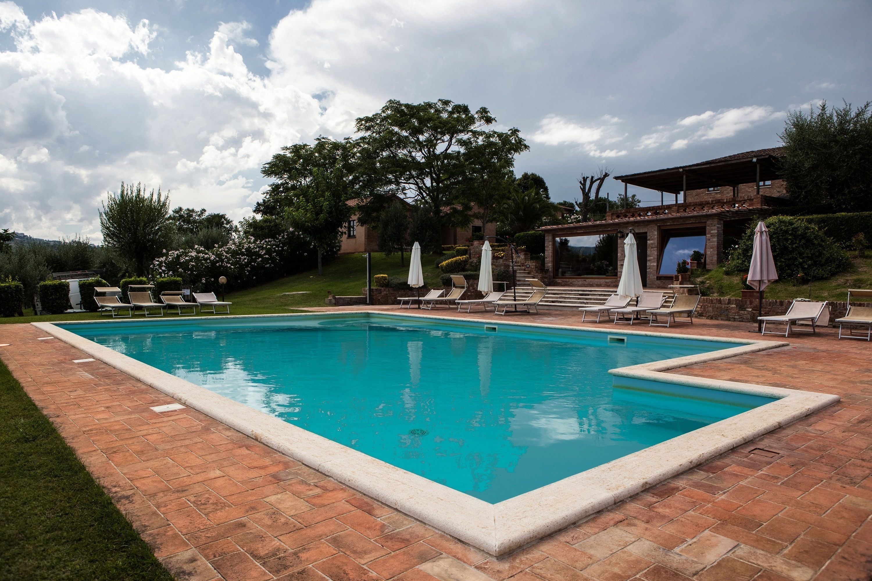 A rectangular swimming pool with a tiled deck, surrounded by lounge chairs and umbrellas, with a building in the background.