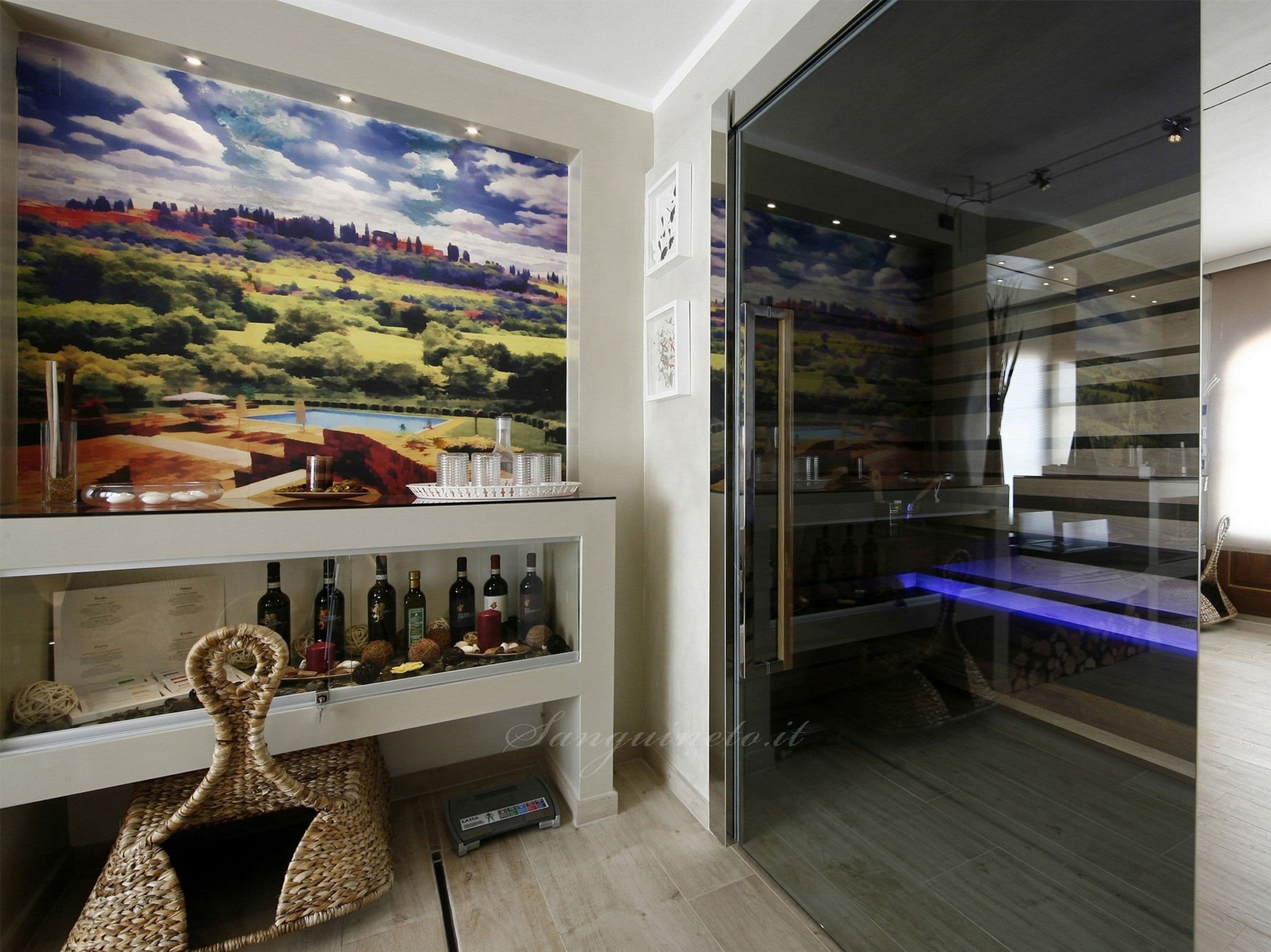 Una cava de vinos con un amplio mural de un paisaje, estantes llenos de botellas y una vitrina de vidrio.