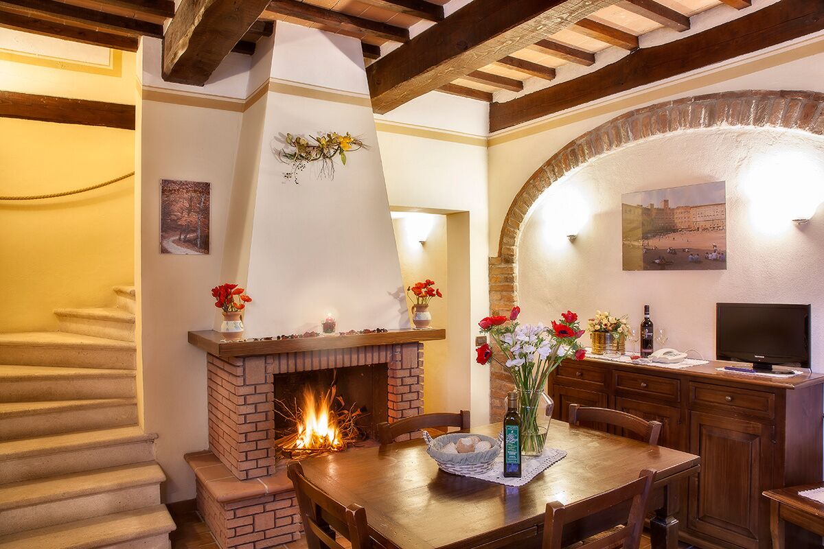 A cozy dining area with a brick fireplace, wooden dining table, and chairs. There is a TV on a wooden cabinet and a vase with flowers on the table.