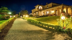 A well-lit, historic building with a gravel pathway leading to the entrance, surrounded by neatly trimmed hedges and flowering plants.