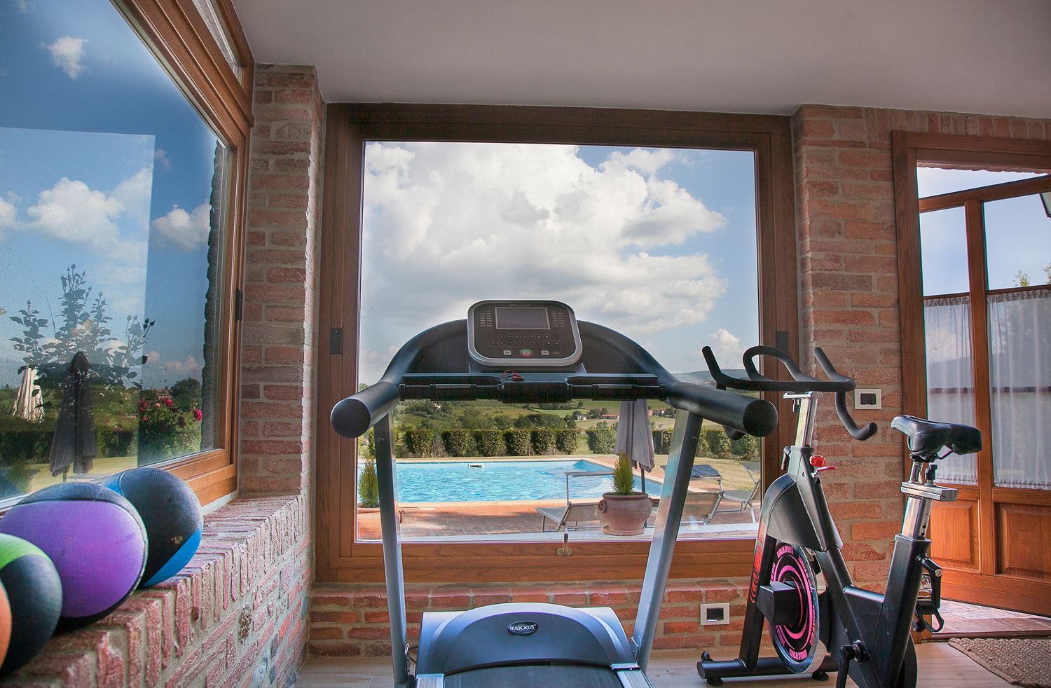 Un gimnasio en casa con equipo de ejercicio, vista a una piscina y un muro de ladrillo.