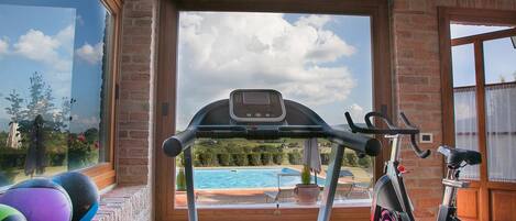Un gimnasio en casa con equipo de ejercicio, vista a una piscina y un muro de ladrillo.