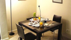 Un comedor con una mesa dispuesta para dos personas, con un frutero, un vaso de jugo de naranja y un florero con una sola flor.