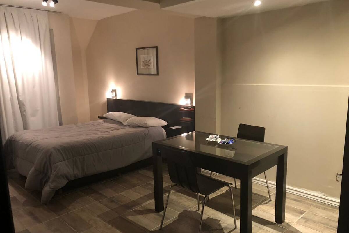 A bedroom with a bed, a desk, and a chair. There is a nightstand with a lamp and a framed picture on the wall.